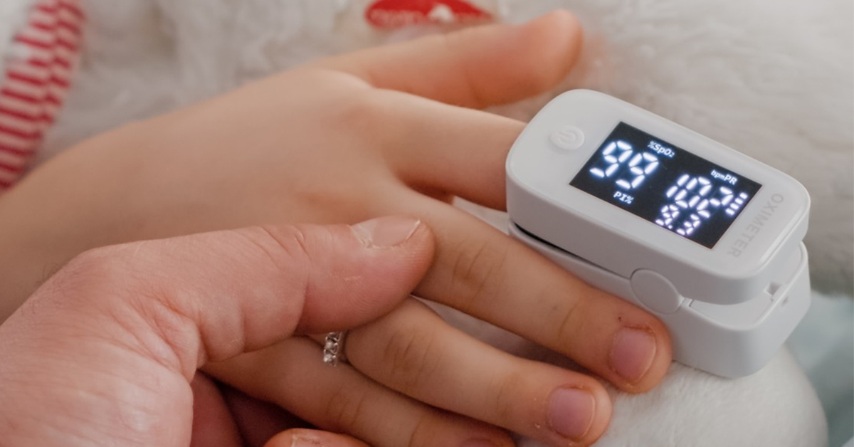 A white pediatric pulse oximeter clipped onto a small child's finger, held by an adult, displaying a healthy blood oxygen level (SpO2) of 99%.