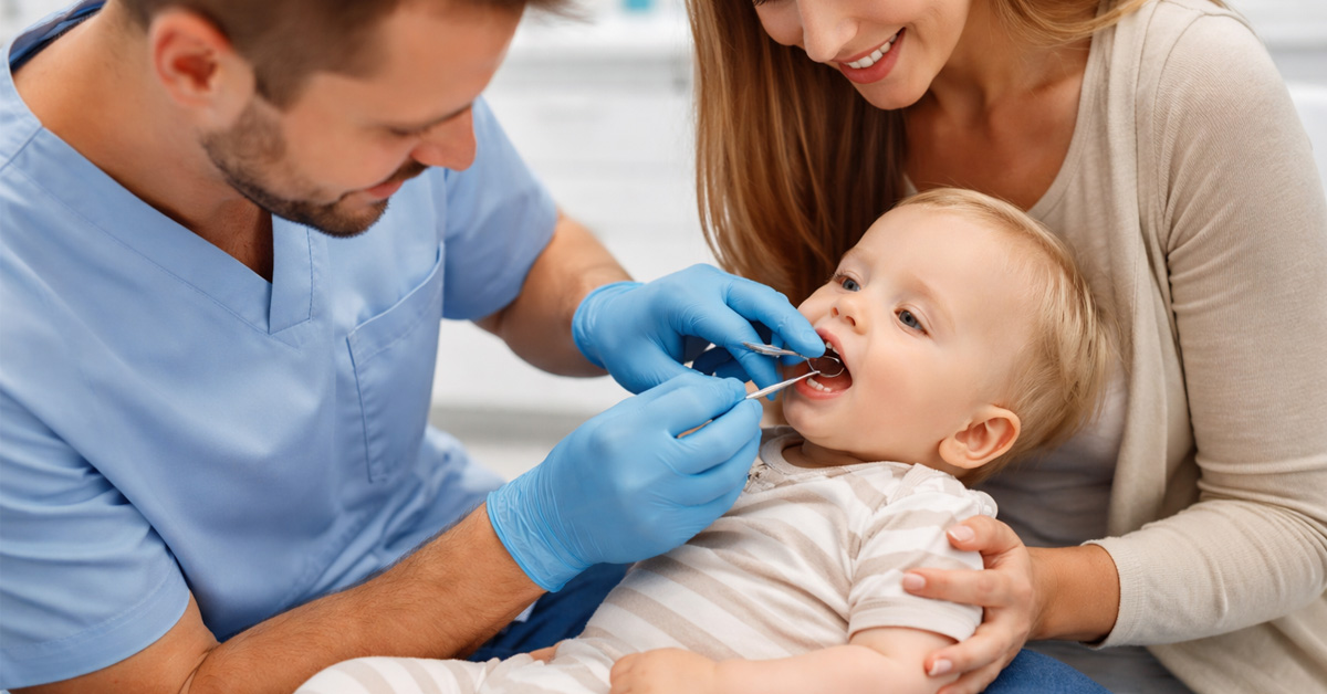 Pediatric dentist examining a baby's teeth using the knee-to-knee method while the parent holds the baby and the mother watches.